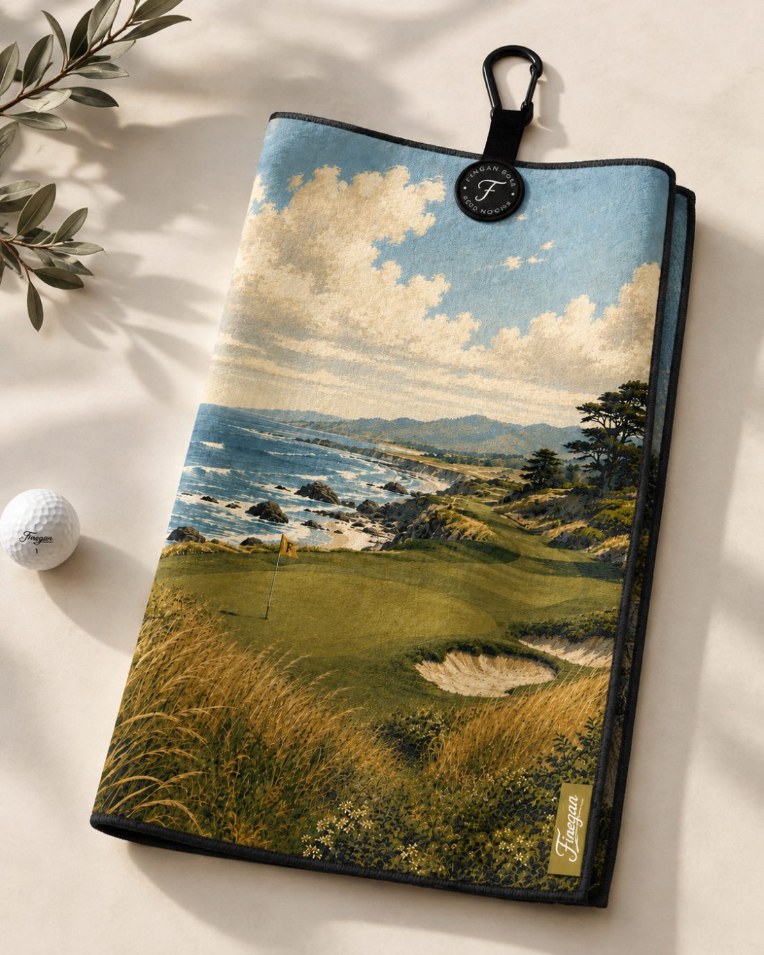 The Cliffs towel folded on a linen surface beside a Finegan golf ball and an olive branch, showing the suede printed face